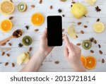Dieting concept. Lady holding cellphone with blank black screen on wooden table with fruit slices, almond and spices, top view, flat lay, mockup