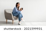 Full length of young woman drinking hot coffee in cozy armchair against white studio wall, banner design with free space. Peaceful lady having relaxing day, chilling on lazy morning