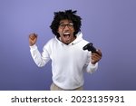 Small photo of Triumphant black teen guy with joystick shouting in excitement, playing video game, making YES gesture on violet background. Overjoyed teenage gamer with controller winning online gaming championship