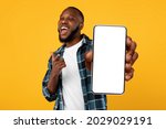 Recommendation. Portrait of excited black guy holding big smartphone with white blank screen in hand, showing close to camera and pointing at device. Gadget with empty free space for mock up, banner