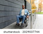 Handicapped accessible city concept. Positive impaired Afro man in wheelchair leaving building on ramp outdoors in autumn, full length. Joyful young black guy using disabled friendly facilities