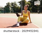 Energy, Vitality, Wellness Concept. Smiling African American woman sitting on basketball field ground, holding shaker, drinking water, taking break after outdoors exercise, wearing wireless earbuds