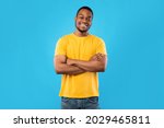 Small photo of Cheerful African American Guy Smiling To Camera Posing Crossing Hands Standing Over Blue Background. Studio Shot Of Happy Self-Confident Black Millennial Man Expressing Positive Emotions