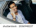 Cheerful young indian man sitting in taxi back seat, talking on cellphone with friend and smiling, shot from window, copy space. Handsome long-haired arab guy using transportation service