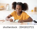 Cheerful inteligent african american teen girl in casual doing homework, sitting at cozy kitchen at home, taking notes or making plan, using pencils and notepad, closeup portrait, copy space