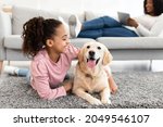 Love And Domestic Animals. Positive happy black teenage girl lying on rug floor carpet and embracing her pet, resting with labrador in living room at home, lady using tablet on couch in background