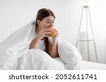 Small photo of Starving upset hungry cute caucasian millennial woman sitting in bed at home and eating burger suffering from stress, depression, crisis and gluttony. Junk food, substitution, diet and too much meal