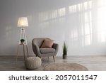 Armchair with pillow, glowing lamp, plant in pot, ottoman and round carpet on floor on gray wall background in living room. Ad blog about real estate and modern interiors, simple scandinavian design
