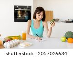 Small photo of Keto diet concept. Happy fit woman writing daily ration diet or menu in notebook, sitting near various products at kitchen table, free space
