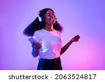 Happy African American woman with headphones and smartphone listening to music and dancing in neon light. Young black woman moving to favorite song, enjoying cool playlist