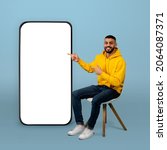 Positive arab guy pointing at big smartphone with white screen while sitting on chair over blue background. Man demonstrating free space for your app or website design, mockup