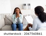 Successful psychotherapy. Millennial female client having session with psychologist, smiling, sitting on couch at clinic. Cheerful black lady communicating with therapist, solving emotional problem