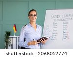 Small photo of Portrait of cheerful German teacher standing near blackboard, conducting internet lesson and smiling at camera. Positive tutor giving language class on video call or web conference