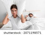 Small photo of Despaired millennial european wife sits on bed covers ears with pillow and suffers from snoring of sleeping husband in bedroom interior. Intolerable snoring and noise at night, insomnia and apnea