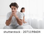 Small photo of Depressed young caucasian husband with stubble sits on bed and sad, ignoring offended lady in bedroom interior. Problems in bed and relationships, male strength and impotence, serious scandal at home