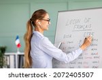 Small photo of Remote education and tutoring concept. Female teacher writing grammar rules on whiteboard during online French lesson, happy tutor teaching foreign language on web