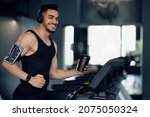 Small photo of Happy Young Middle Eastern Male Athlete Jogging On Treadmill At Gym, Cheerful Muscular Arab Man Holding Sport Shaker Bottle And Wearing Wireless Headphones, Listening Music While Training, Copy Space