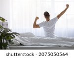Rear View Of Young Man Stretching In Bed After Waking Up In The Morning, Unrecognizable Male Resting In Light Bedroom After Good Sleep, Looking At Window, Enjoying Start Of New Day, Copy Space