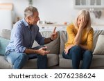 Small photo of Family Conflict. Angry Senior Husband Shouting At Offended Wife Having Quarrel Sitting On Sofa At Home. Marital Problems, Marriage And Relationship Issue Concept