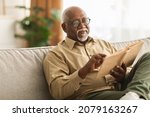 Senior African American Man Reading Book Sitting On Couch At Home, Wearing Eyeglasses. Retired Male Enjoying Reading New Novel Or Business Literature On Weekend. Retirement Leisure Concept