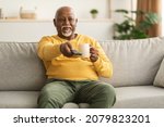 Small photo of Contented Senior African American Man Watching TV Pointing Television Remote Control To Camera Switching Channels Sitting On Couch At Home. Male Viewer Enjoying Movie On Weekend