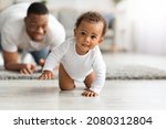 Small photo of Cute Little Black Infant Baby Crawling On Floor At Home, Proud Young Father Looking At Him And Smiling, Dad And Toddler Child Enjoying Spending Time Together, Selective Focus With Copy Space