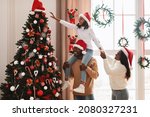 Happy loving family decorating Christmas tree. Young black man holding his daughter on shoulders helping decorate Xmas tree hanging Santa Claus hat on top, people standing in living room together
