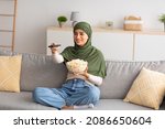 Portrait of young Arab female in hijab watching TV with remote control and popcorn, sitting cross legged on couch at home. Middle Eastern lady in headscarf enjoying good movie with yummy snack