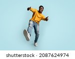 Small photo of Carefree african american young man in casual outfit showing thumb ups and raising leg up, happy black guy posing on blue studio background, full length shot, panorama with copy space