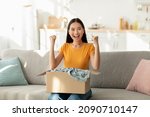 Small photo of Overjoyed asian woman unboxing cardboard box parcel, emotionally reacting to successful shopping, clenching fists with excitement, sitting on couch in living room