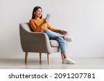 Cheerful millennial Asian woman using mobile phone, chatting on web, working or studying online, sitting in armchair against white wall, copy space. Pretty young lady watching video on smartphone