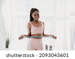 Portrait of cheerful young African American lady in sportswear measuring waist with tape, feeling happy about losing weight at home. Healthy lifestyle, sports and slimming diet concept