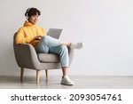 Happy young Asian man using laptop, wearing headphones while sitting in armchair against white wall, copy space. Millennial male communicating online, working or studying remotely on portable pc