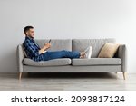 Cool Gadget And Application. Portrait of young smiling Arab man holding mobile phone, typing sms message, sitting on the sofa in living room. Guy browsing internet, surfing web, using app, free space