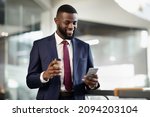 Cheerful young black bearded man manager standing at office building corridor, drinking coffee to go, using mobile phone, chatting with business partner and smiling, panorama with copy space