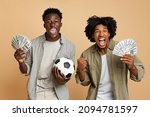 Sport Bets. Portrait Of Excited Black Guys With Football Ball And Money Standing Over Beige Background, Cheerful Soccer Fans Celebrating Success With Dollar Cash, Emotionally Reacting To Win