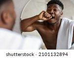 Sleepy Black Guy Yawning Holding Fist Near Mouth Standing Shirtless Looking At His Reflection In Mirror In Modern Bathroom At Home In The Morning. Male Beauty Routine Concept. Selective Focus