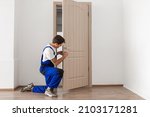 Small photo of Maintenance Concept. Focused serious young locksmith with tattoo on hand wearing blue uniform standing on one knee in doorway installing doorknob handle, repairing wooden front door entrance