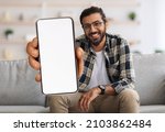 Cheerful Indian Guy Showing Smartphone With Big Blank White Screen At Camera, Happy Young Eastern Man Recommending New Mobile App Or Website While Sitting On Couch At Home, Creative Collage, Mockup