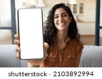 Mobile Advertisement. Happy Beautiful Woman Showing Big Blank Smartphone At Camera While Sitting On Couch At Home, Young Female Demonstrating Phone With White Screen For App Design, Collage, Mockup