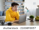 Bored young Asian guy sitting at desk using pc laptop, looking aside, thinking about something during online lesson or remote work at home office. Tired man having dull distant job, feeling lazy