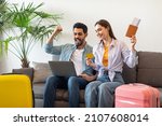 Small photo of Excited tourists couple booking hotel room online, using laptop and credit card, sitting on sofa with suitcases, passports and tickets, ready for vacation trip