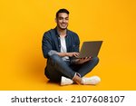 Happy arab guy in casual freelancer using modern laptop, working online, yellow studio background. Cheerful middle eastern young man independent contractor sitting on floor with notebook, copy space