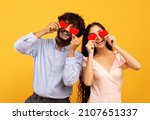 Lovers blinded by their big love. Young indian couple in love holding red heart-shaped cards over eyes and smiling, standing over yellow studio background. St. Valentines Day celebration concept