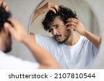 Small photo of Men's beauty and haircare treatment. Worried indian guy looking at flaky scalp in mirror, examining gray hair and hairloss issue, standing in bathroom at home.
