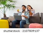 Small photo of Happy young couple using laptop and booking hotel online with credit card, woman holding passports with tickets, sitting with packed suitcases on sofa at home