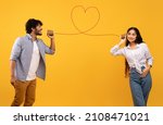 Love message. Loving indian man telling romantic words to girlfriend through tin can phone with heart shaped string, standing over yellow background, creative collage