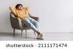 Small photo of Peaceful young Asian man relaxing in cozy armchair, being lazy, having break against white studio wall, panorama with free space. Handsome millennial guy resting with hands behind head