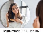 Haircare Concept. Attractive Young Lady Brushing Her Thick Beautiful Hair With Comb While Standing Near Mirror In Bathroom, Happy Woman Wearing White Silk Robe Looking To Her Reflection And Smiling