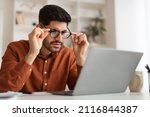 Small photo of Error. Portrait of confused Arab guy sitting at desk using laptop, taking off glasses looking at pc screen and squinting. Worried man reading bad negative news, having poor eyesight problems